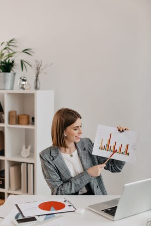 Young business woman in white office with smile shows graphic, talking to customers on Skype.